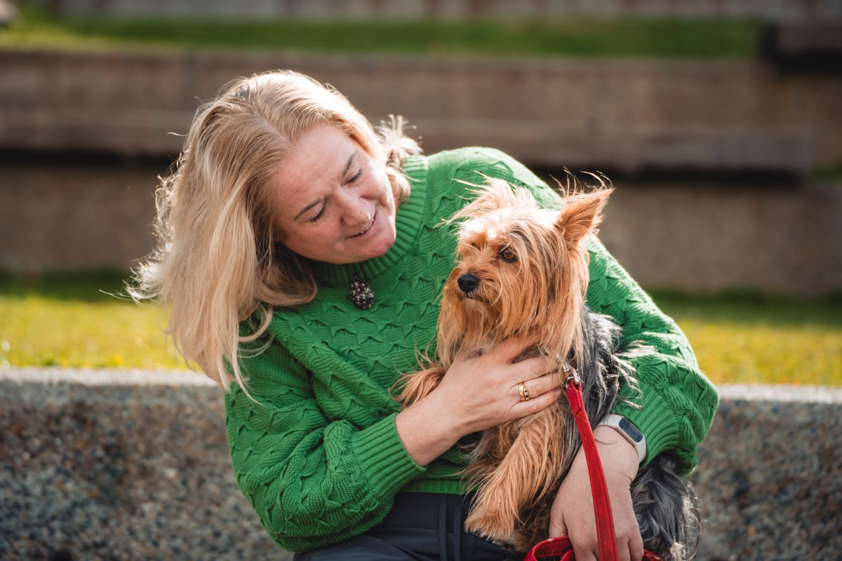 Woman smiling at a small dog on her lap outdoors.