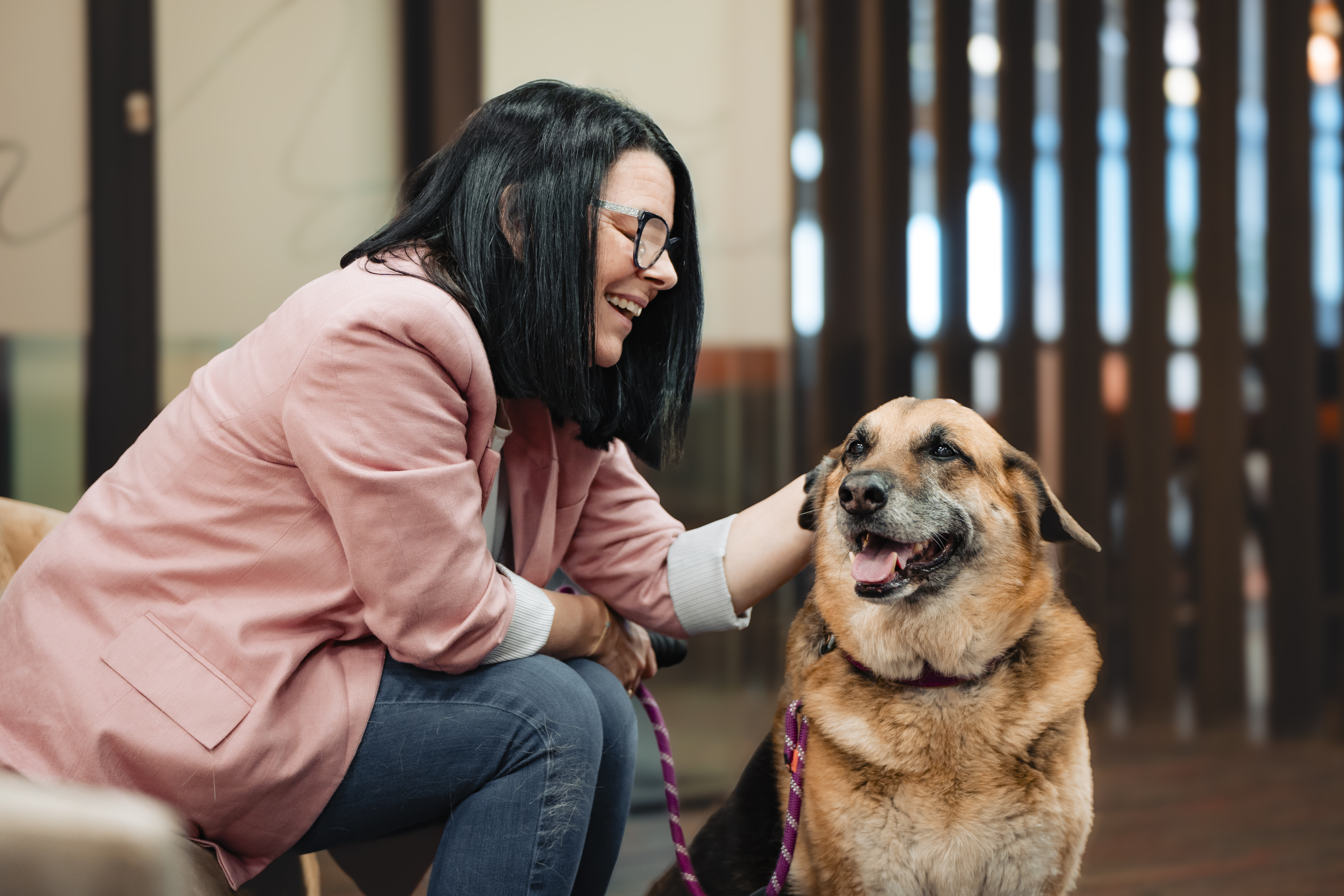 Woman in pink blazer smiling at a dog indoors.