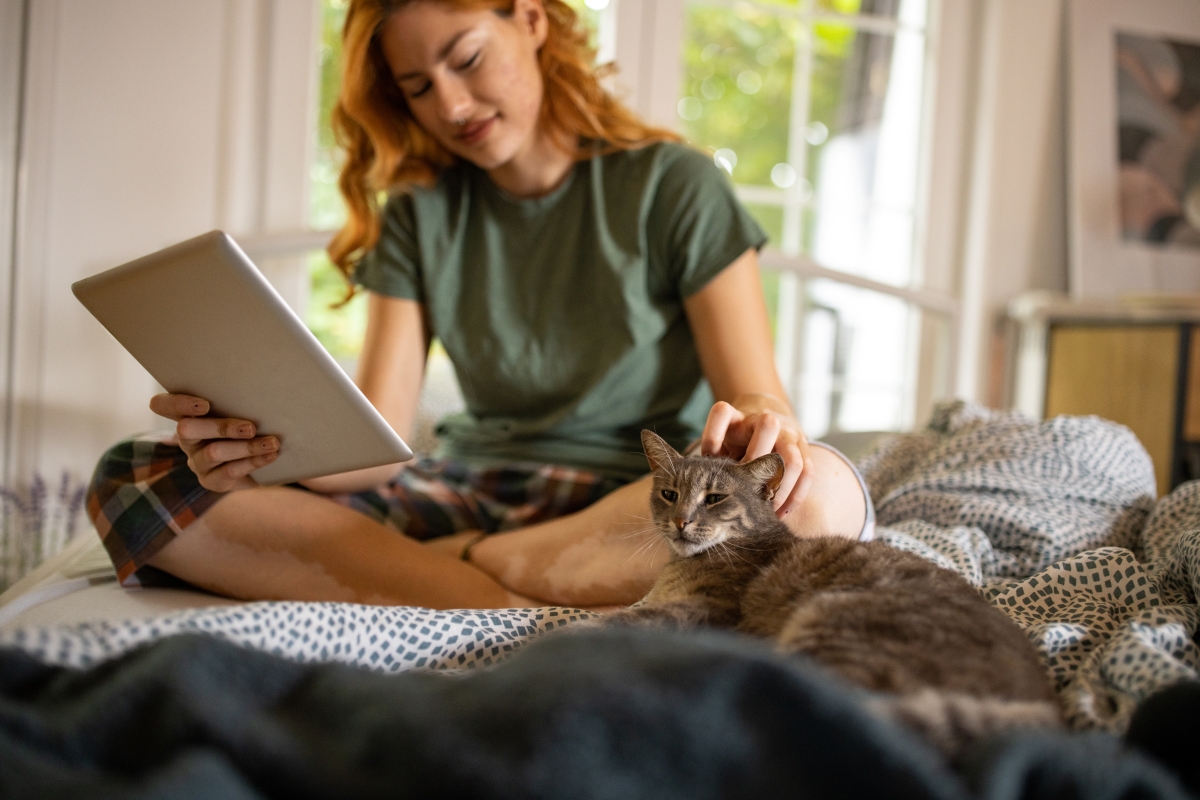 Woman petting a cat while using a tablet on a bed.