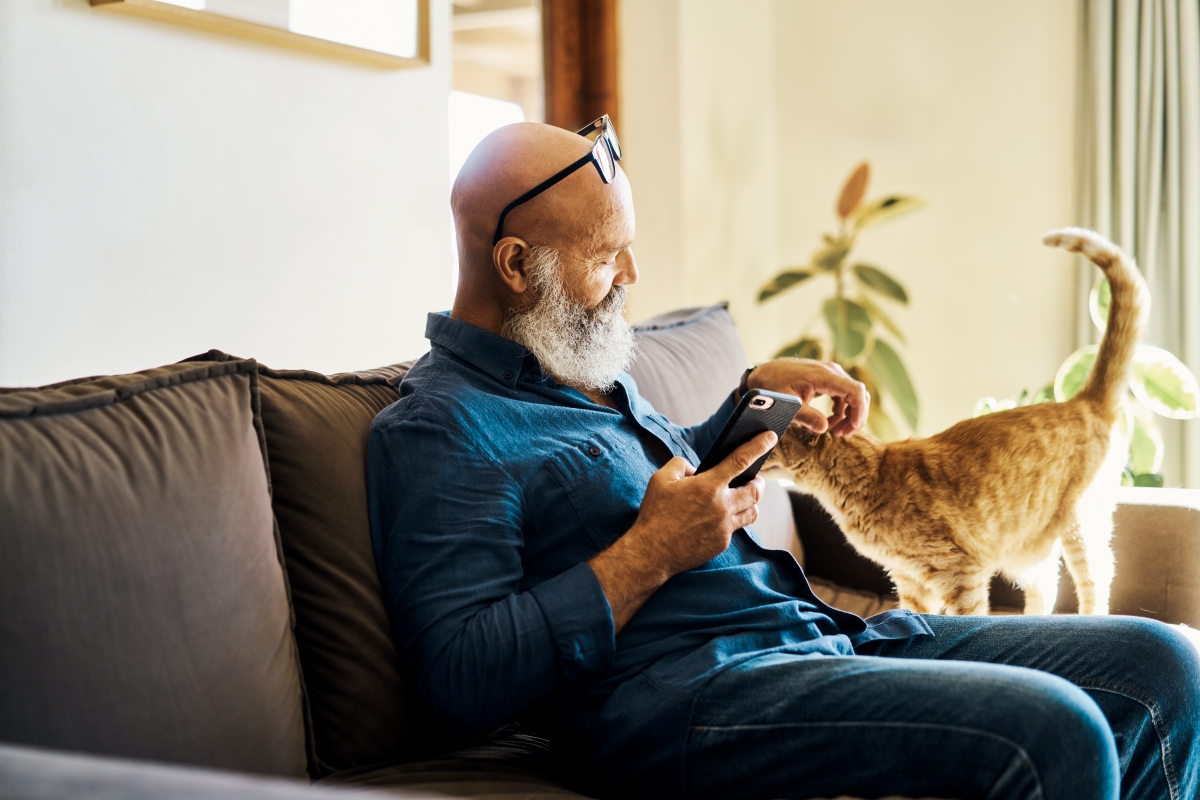 Man petting a cat while holding a phone on a sofa.
