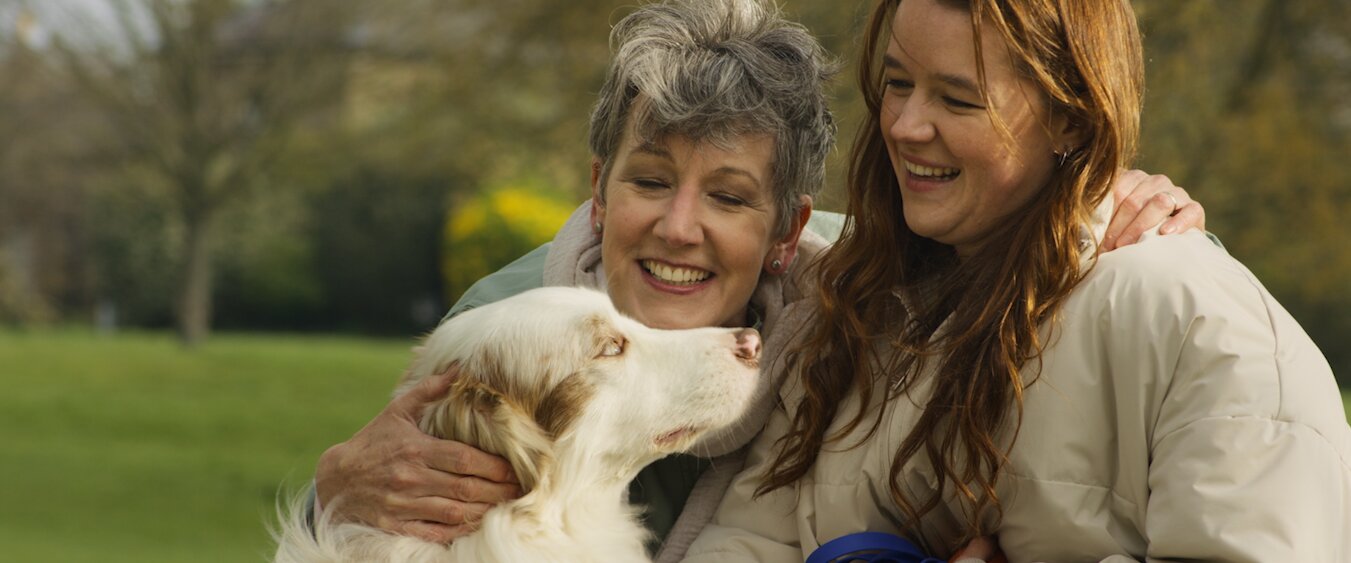 Two women smiling at a dog in a park.